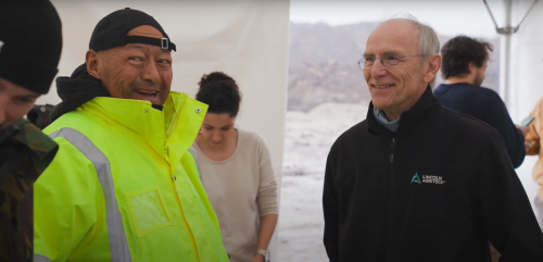 A man wearing a yellow forestry safety jacket stands next to an older man wearing a navy Lincoln Agritech jersey inside a white weather gazebo.