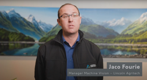 A man wearing glasses stands before a large image of New Zealand mountain scenery